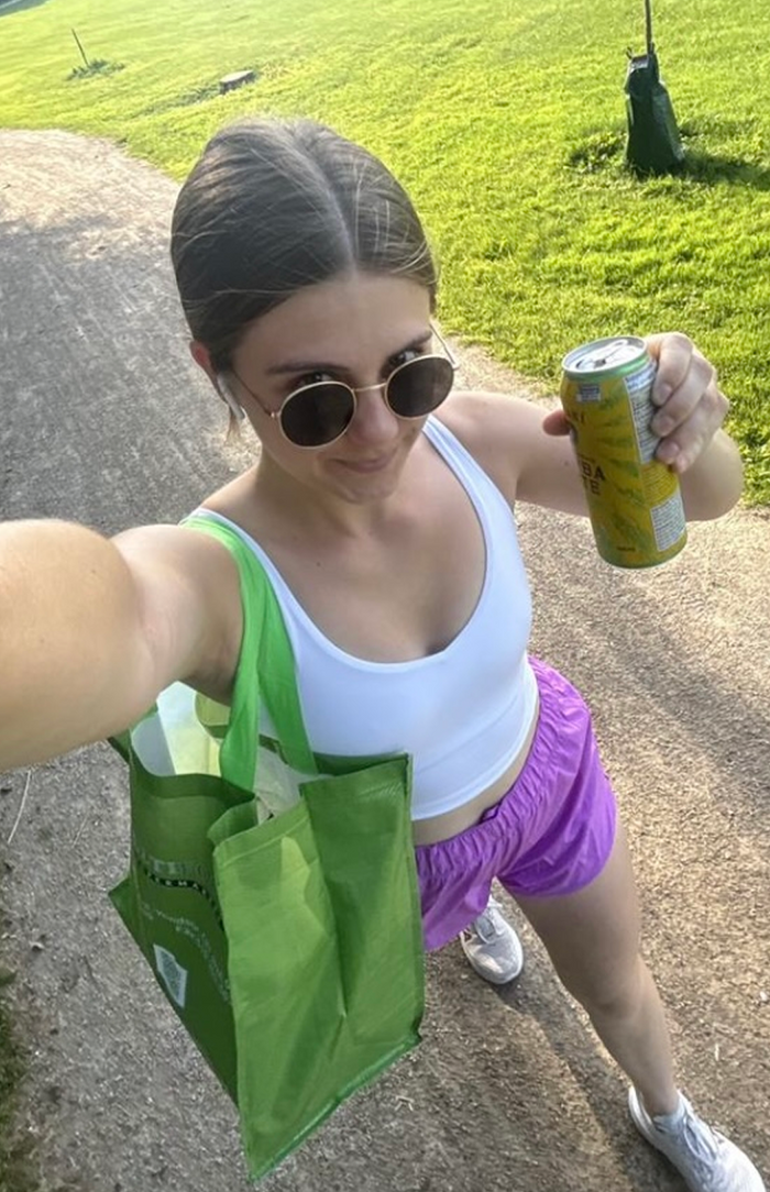 A woman taking a selfie while walking on a sunny path, wearing sunglasses, a white crop top, and purple shorts, holding a can of Yerba Madre in one hand and a green tote bag in the other.