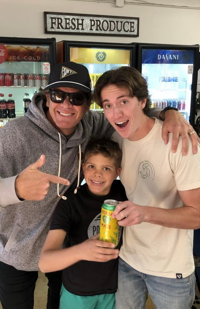Three smiling people pose inside a store in front of refrigerators, with one child holding a can of Yerba Madre as the others point and smile at the camera.