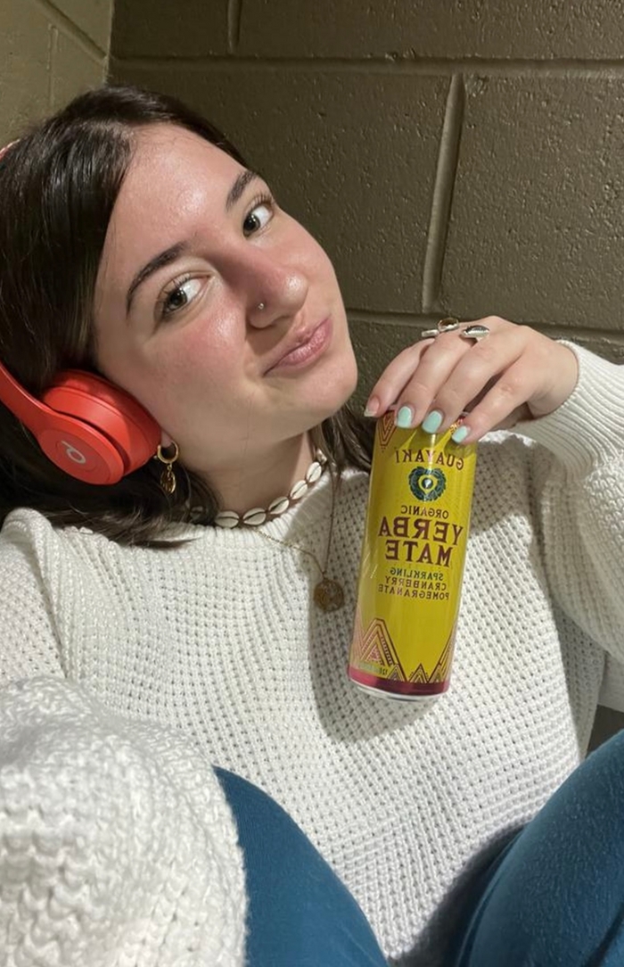A young woman wearing orange headphones and a white sweater smiles at the camera while holding a can of Yerba Madre, sitting against a tan brick wall.
