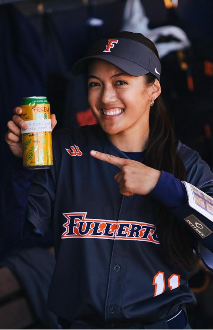 A smiling softball player in a Fullerton uniform and visor points to a can of Yerba Madre, proudly holding it up in a dugout setting.