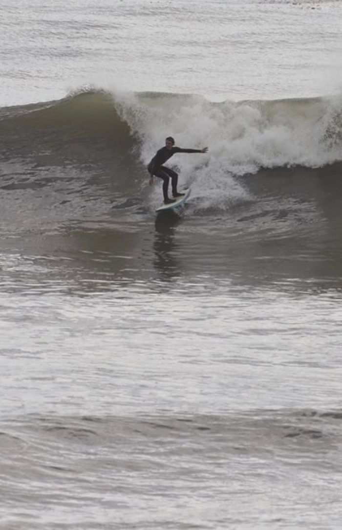 A surfer in a wetsuit rides a breaking wave on a cloudy day, balancing on the board as the wave curls behind them.