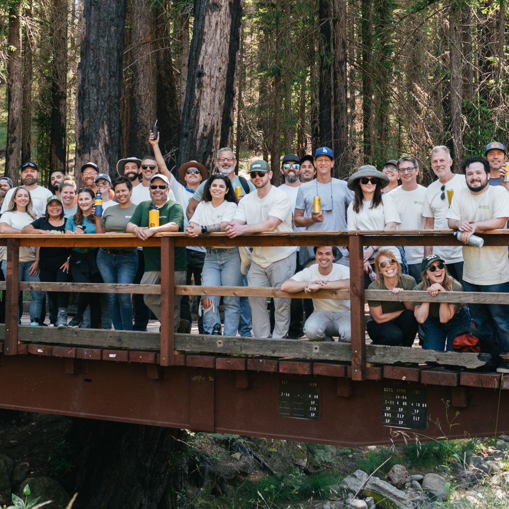 A large group of people stand together on a wooden bridge in a forest, smiling and holding cans of Guayakí Yerba Mate. They are dressed casually, with some wearing hats and sunglasses, surrounded by tall trees.
