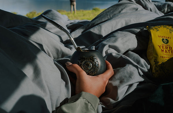 Person lying under gray blankets outdoors, holding a traditional mate gourd with a metal straw, next to a bag of Yerba Madre, with a blurred lake and figure in the background.