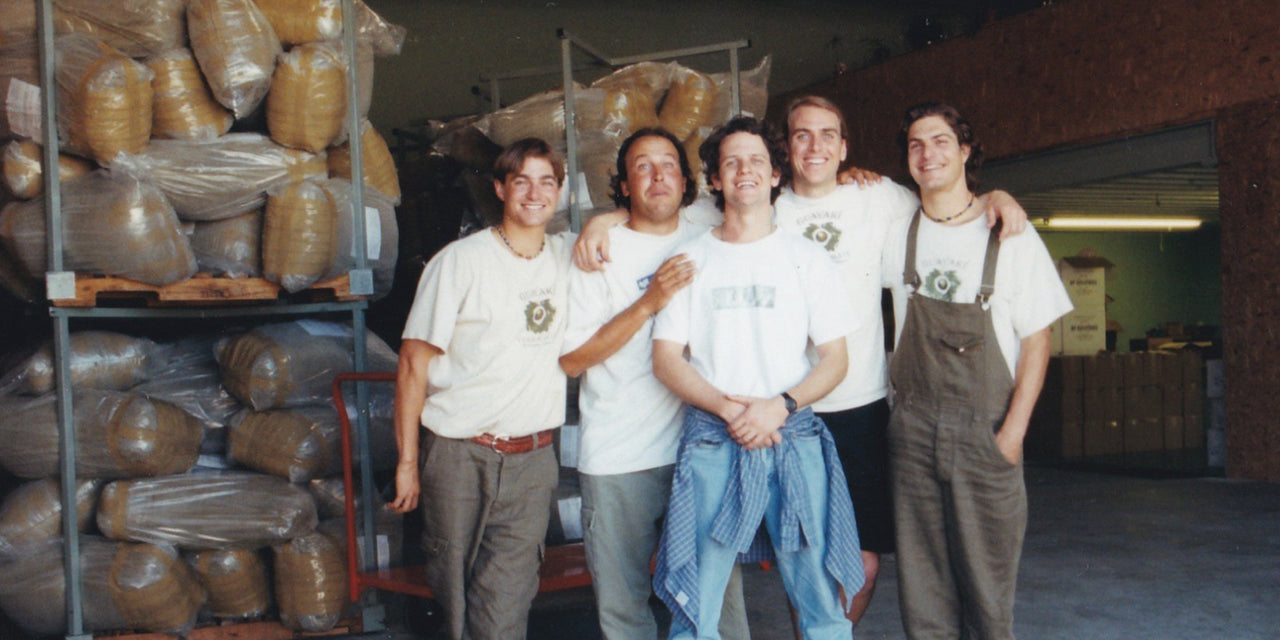 Five people stand smiling in a warehouse filled with large bags of organic products on shelves behind them. Casually dressed, with some in t-shirts and one in overalls, they exude the relaxed and cheerful energy of a team proud to craft the high-energy cans of Guayakí Yerba Mate.
