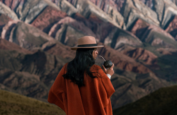 A woman in a wide-brimmed hat and rust-colored poncho stands facing colorful, layered mountains, sipping from a traditional mate gourd with a metal straw.