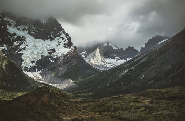 Dramatic view of a remote mountain valley surrounded by steep, jagged peaks with patches of snow and glaciers under heavy, moody clouds.